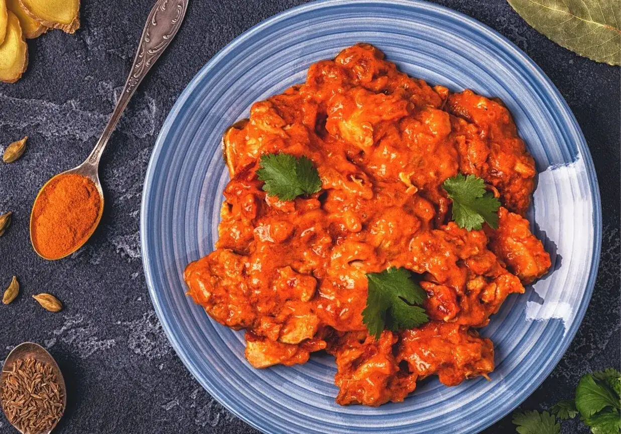 Aksamitny chicken masala na niebieskim talerzu, udekorowany kolendrą. Obok łyżeczki z kurkumą i kminem.