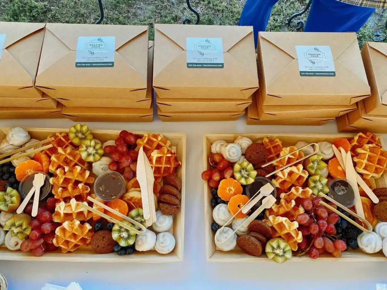 Dwa drewniane tace pełne pyszności: gofry, owoce (winogrona, kiwi, mandarynki, borówki), ciasteczka i słodkie bułeczki. Idealne dietetyczne śniadanie na wynos.