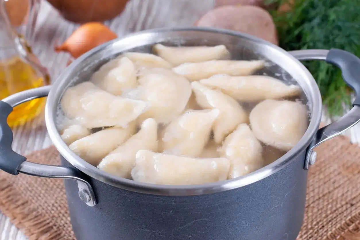 Pierogi gotują się w garnku. W tle olej, cebula i koperek. Dowiedz się, ile gotować pierogi, by były idealne.