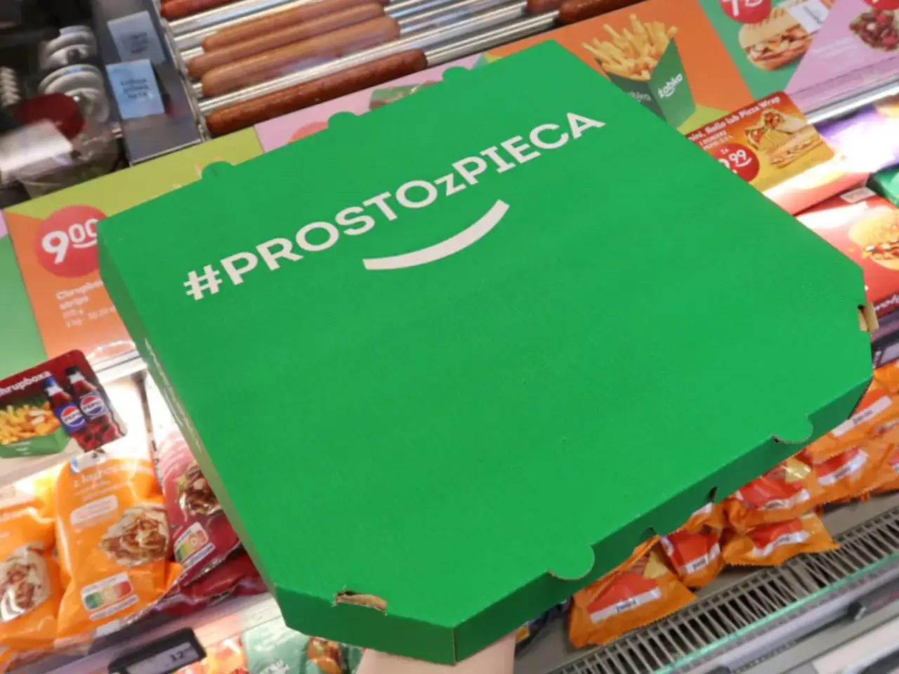 Zielone pudełko z napisem #PROSTOzPIECA, idealne na pizzę z Żabki. W tle widać inne przekąski i hot dogi.