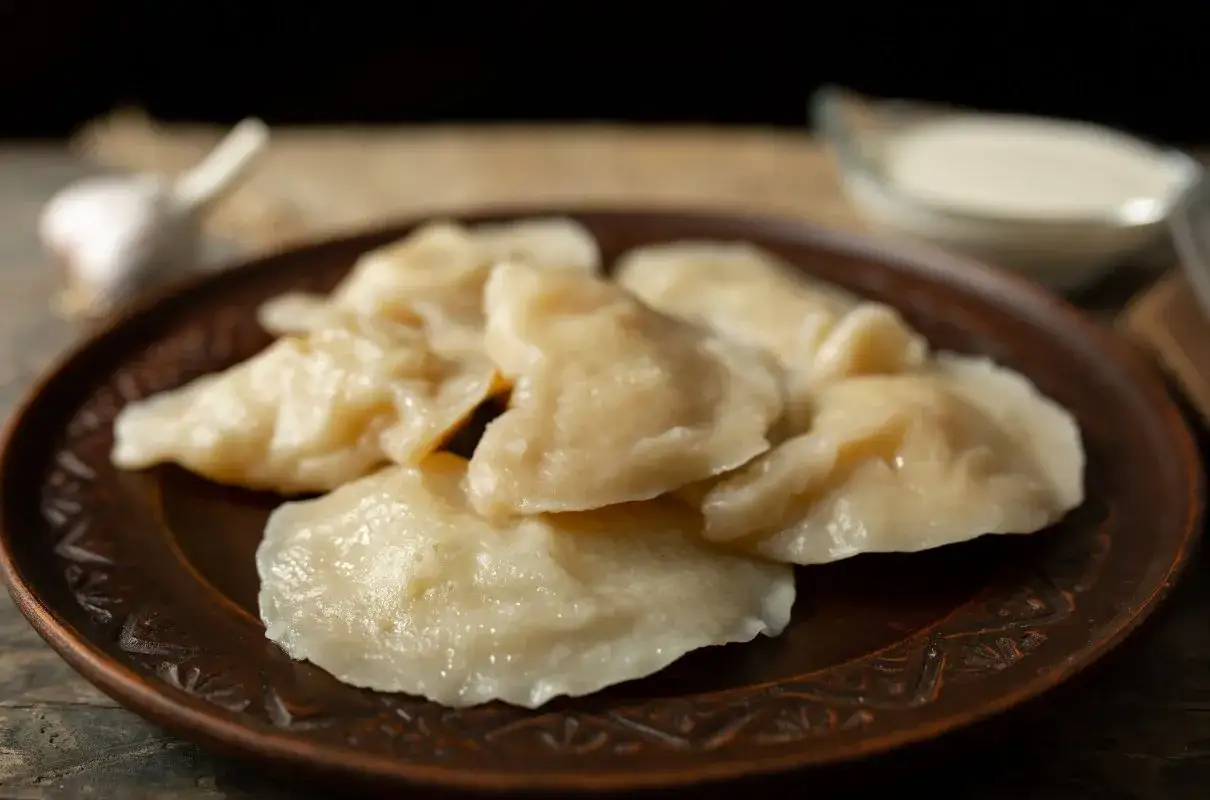Na glinianym talerzu ułożone są pyszne, domowe pierogi chłopskie. Obok czosnek i miseczka śmietany.