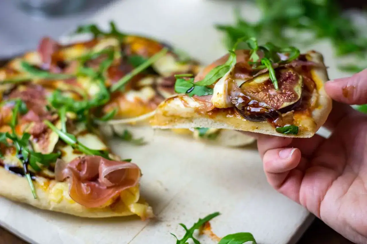 Kawałek pysznej pizzy parma z figami, rukolą i szynką prosciutto, podniesiony do degustacji.