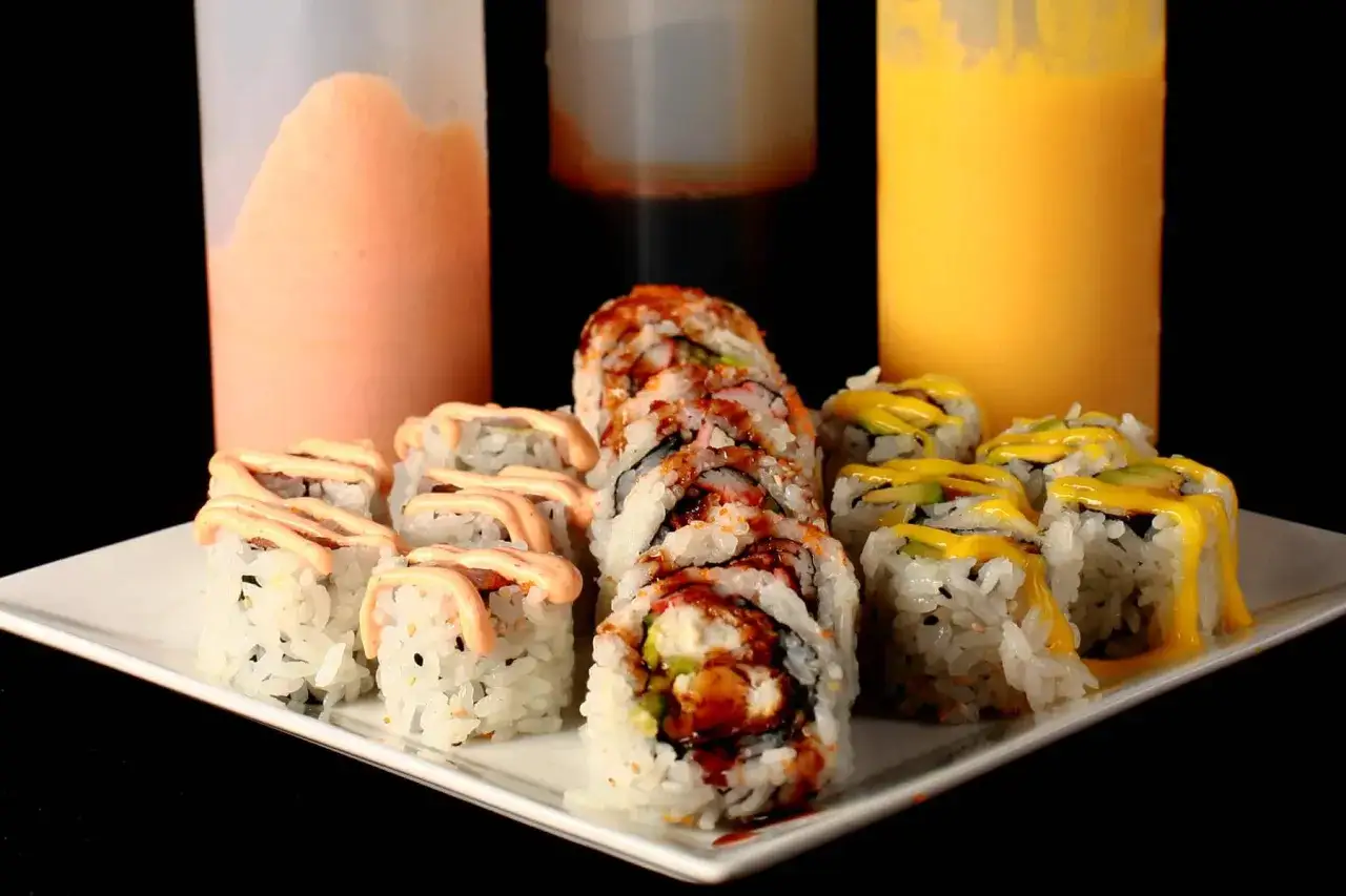 Talerz sushi z różnymi rolkami, polany kremowym sosem do sushi. Obok stoją butelki z pomarańczowym i żółtym sosem.