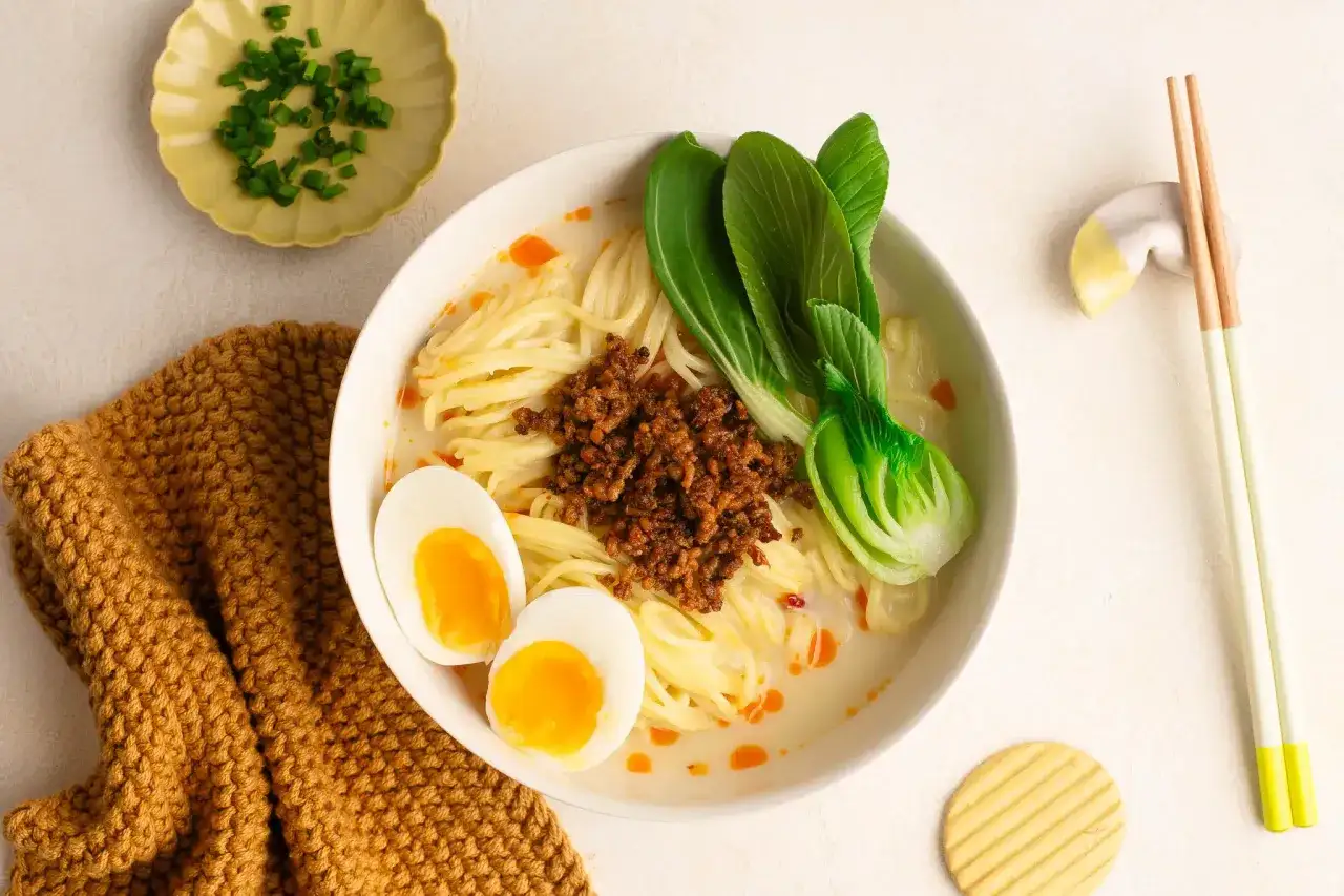 Pyszny tantanmen z mielonym mięsem, jajkiem na pół i pak choi. Obok miski leży pomarańczowy, dzianinowy pled.