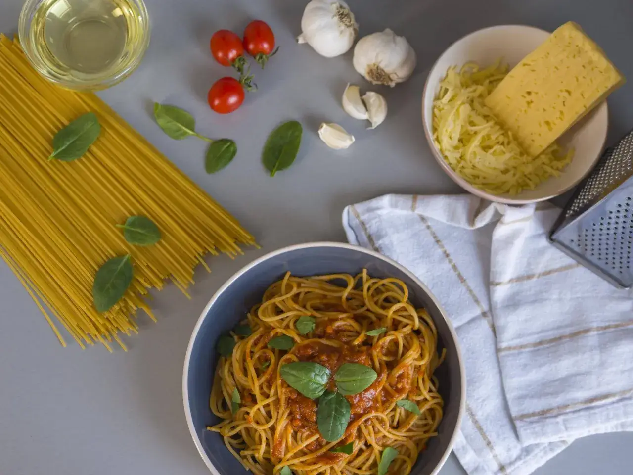 Gotowi na pyszne spaghetti? Na stole składniki: makaron, pomidorki, czosnek, oliwa, ser i bazylia.