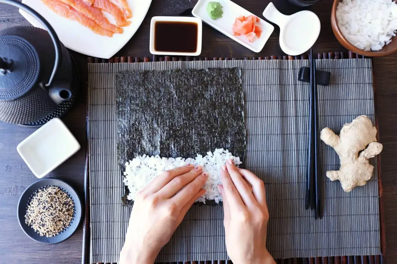 Ręce rozprowadzają ryż na nori, przygotowując zaprawę do sushi. Obok łosoś, sos sojowy, imbir i czarny sezam.