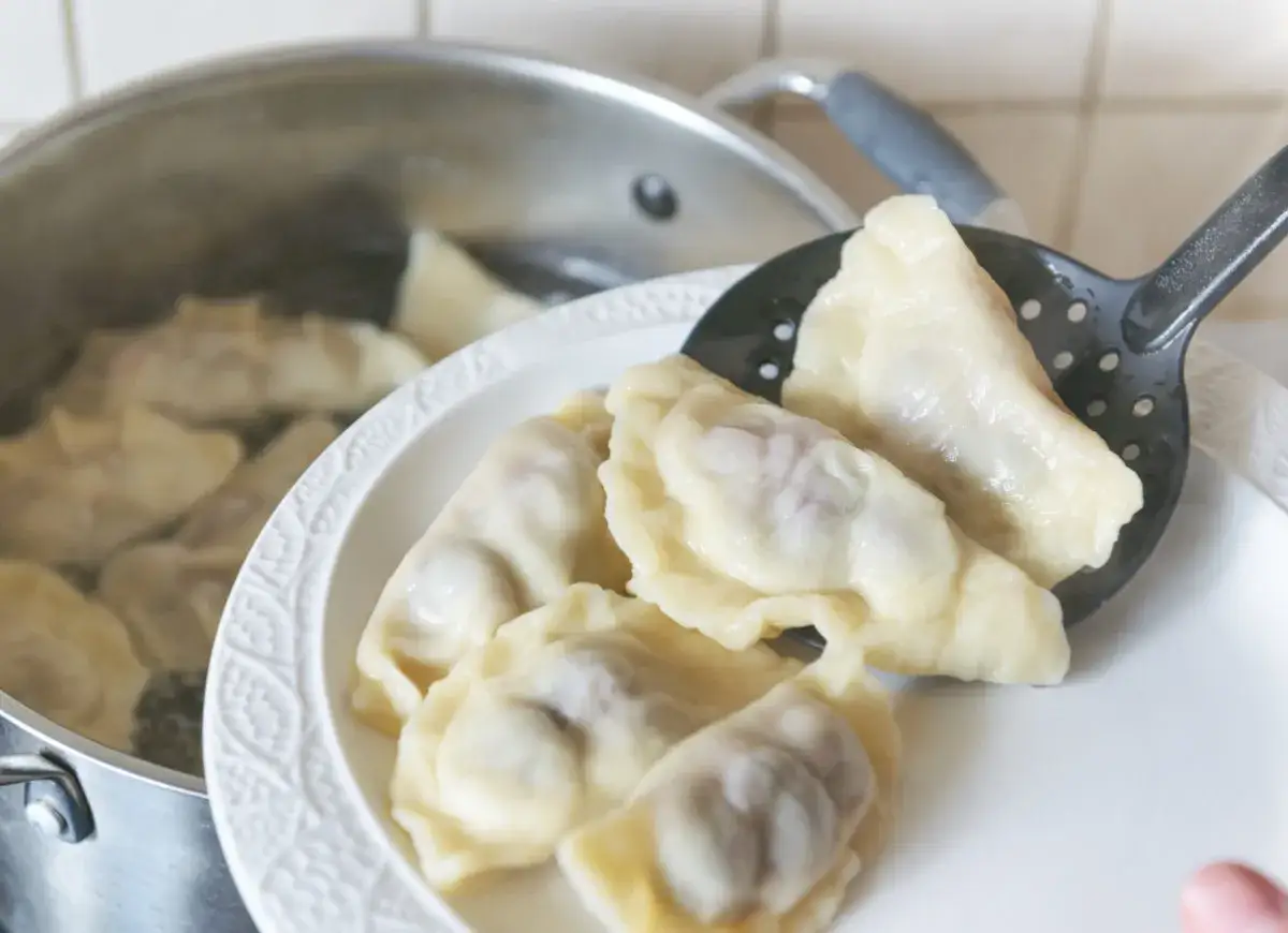Gotowanie pierogów: wyjmowanie łyżką cedzakową gotowych pierogów z garnka na biały talerz.