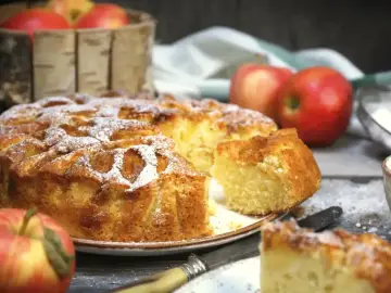 Wilgotne ciasto z jabłkami na oleju, posypane cukrem pudrem, wygląda apetycznie. Obok koszyk z jabłkami i kawałki ciasta.