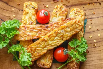 Usmażony tempeh z pomidorkami koktajlowymi i natką pietruszki na drewnianej desce.