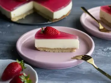 Pyszny deser z mascarpone i galaretką, udekorowany truskawką, na różowym talerzyku. Obok kawałek ciasta i widelczyk.